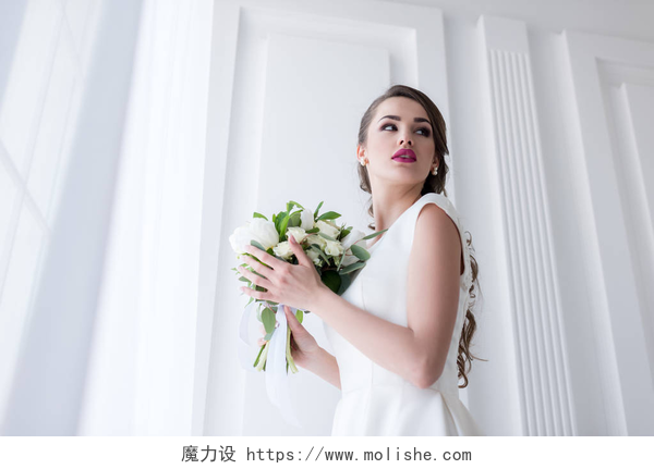 白色墙壁前的一个美丽新娘年轻新娘摆在传统的白色礼服与婚礼花束